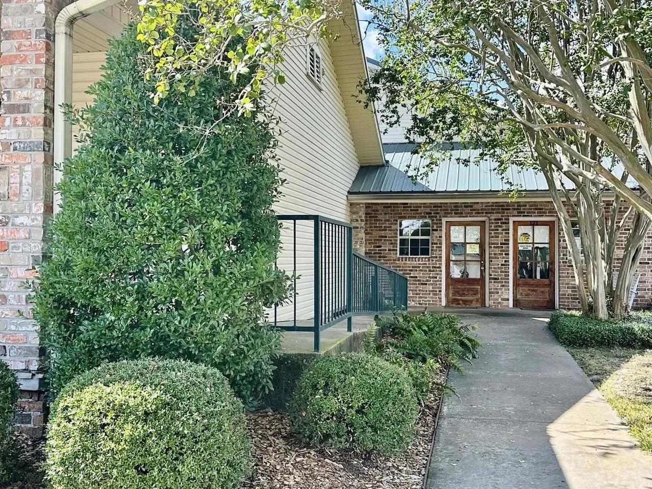 Brick building entrance with walkway, shrubs, and large trees overhead