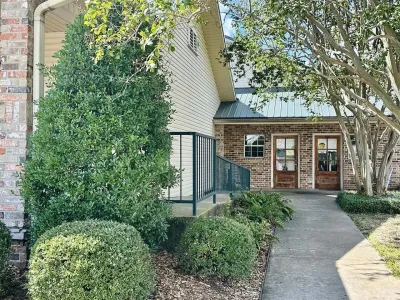 Brick building entrance with walkway, shrubs, and large trees overhead