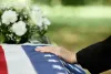 Hand resting on a red, white, and blue-draped casket with white flowers in the background