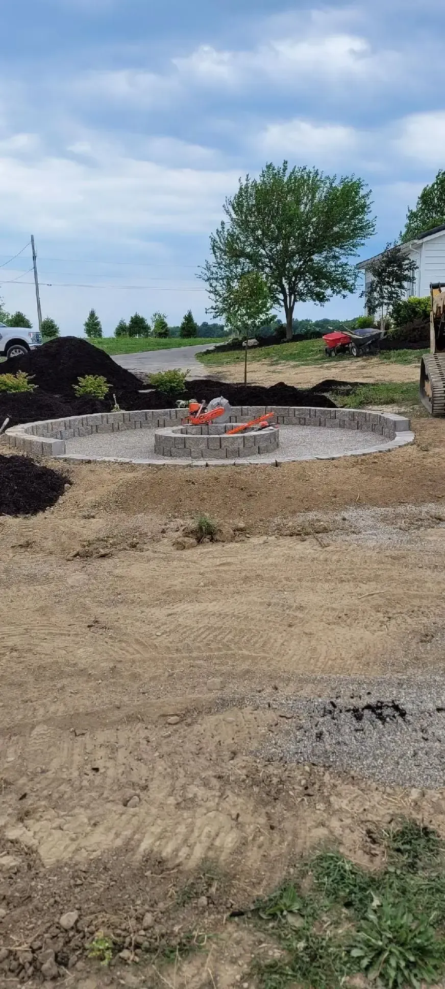 Construction site with gravel, dirt piles, a small stone fire pit, and a tree under a blue sky.