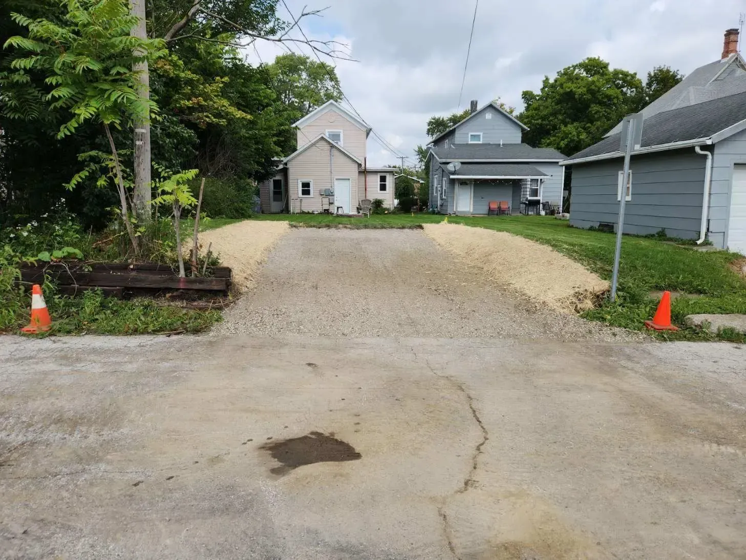 Gravel driveway between houses with orange cones and a small landscaped bed on the left.