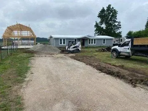 Construction site with gravel road, house under construction on left, and trucks beside a finished home under cloudy sky