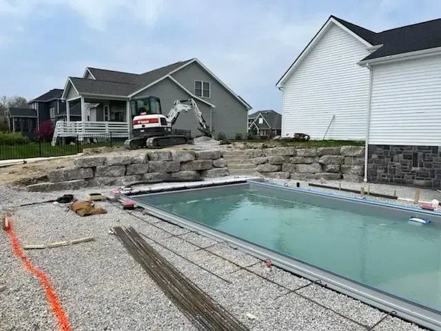 Backyard with an in-ground pool under construction beside a gray house and stone retaining wall.