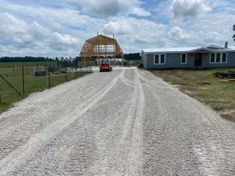 Gravel road leading to a wooden house under construction, with a red car and a cloudy sky