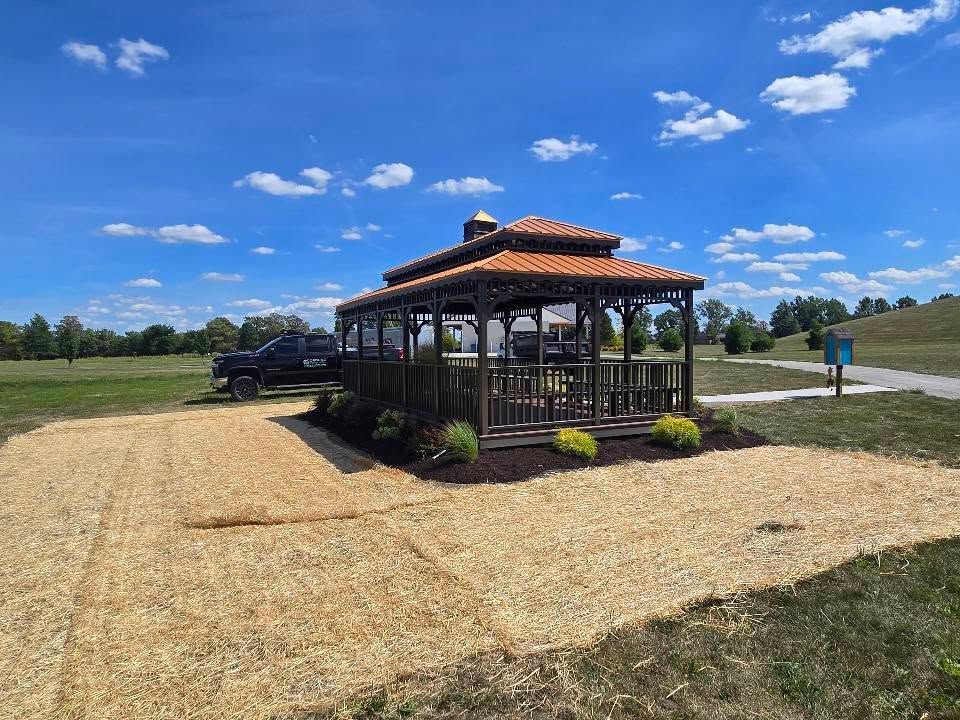 Gazebo with orange roof in a grassy field under a bright blue sky, with parked cars nearby