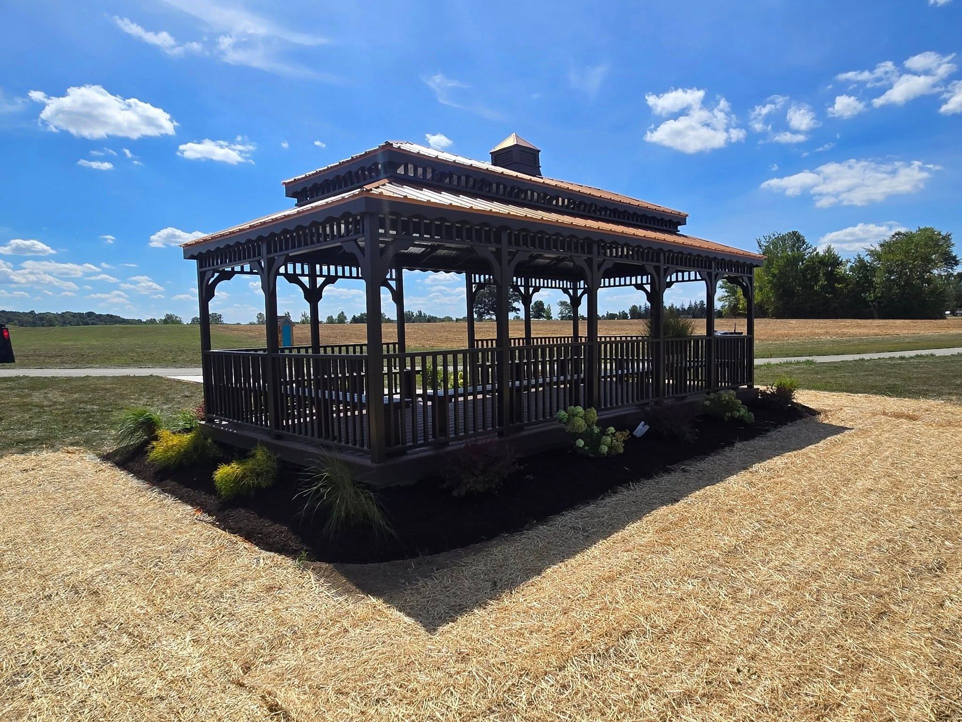 Dark wooden gazebo in a gravel clearing under a blue sky with clouds, surrounded by open fields.