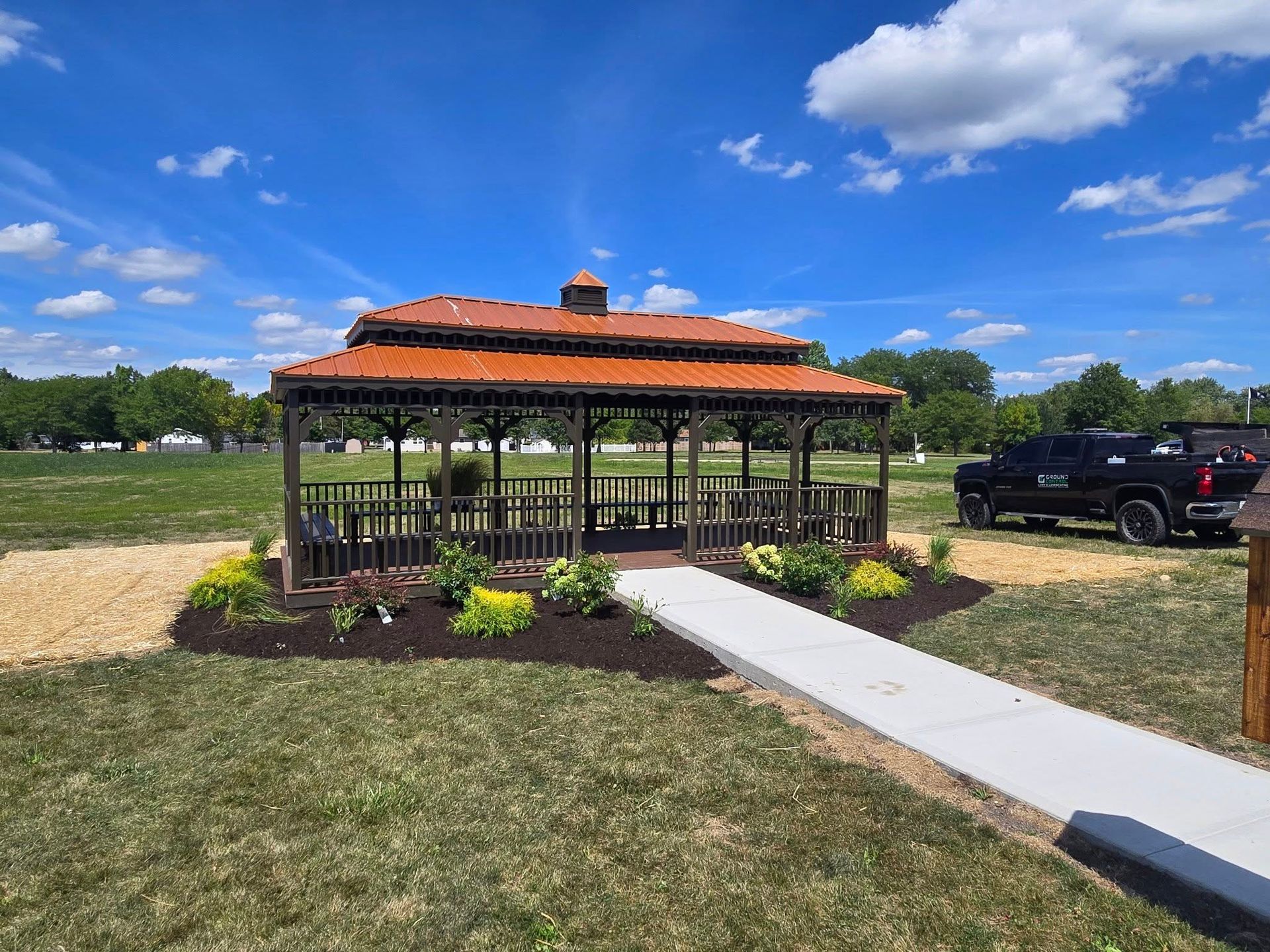 Outdoor gazebo with orange roof beside a sidewalk and parked vehicles in a grassy field under a blue sky