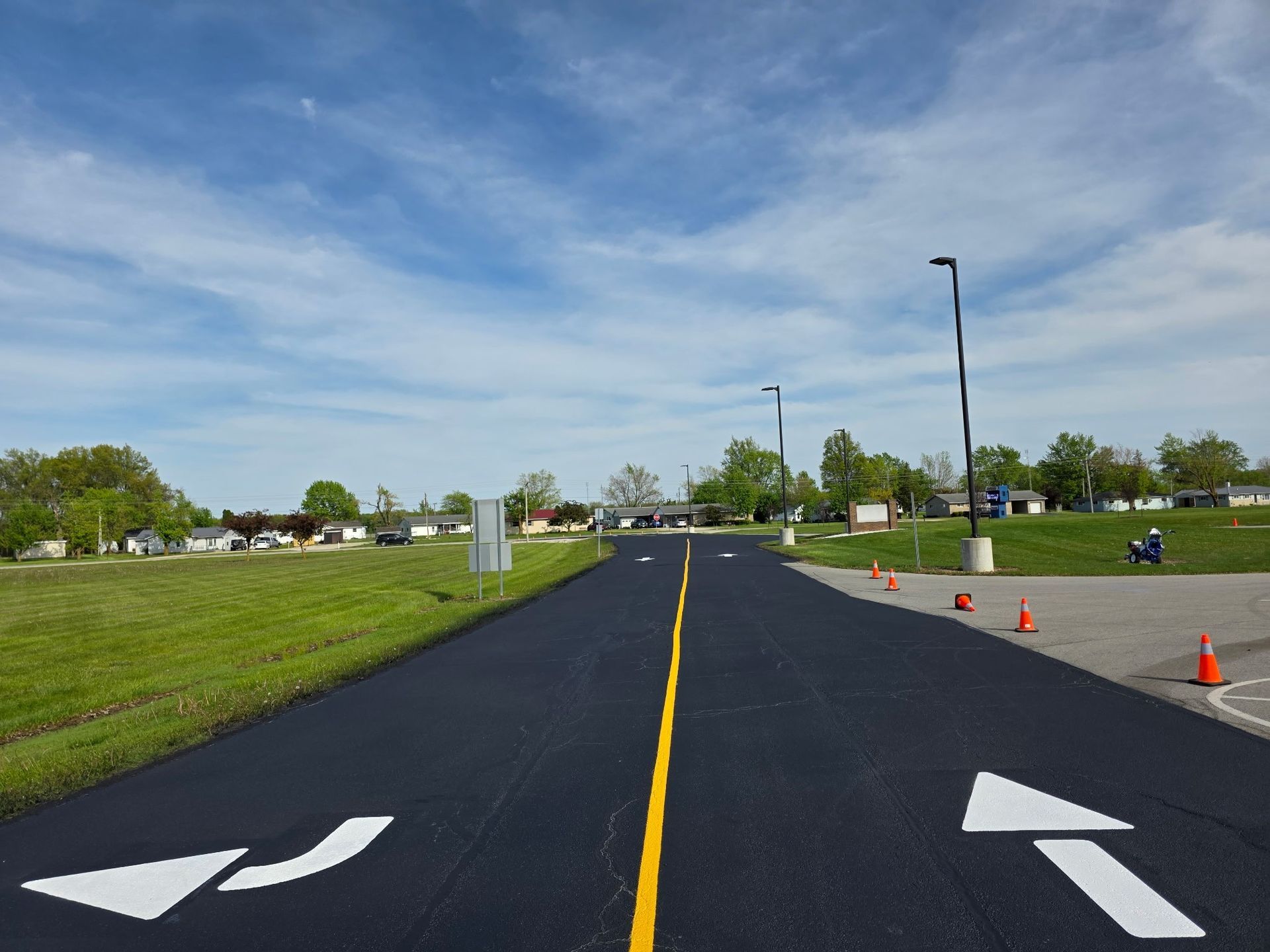 Freshly paved road with white arrows, orange cones, and grassy roadside under a blue sky