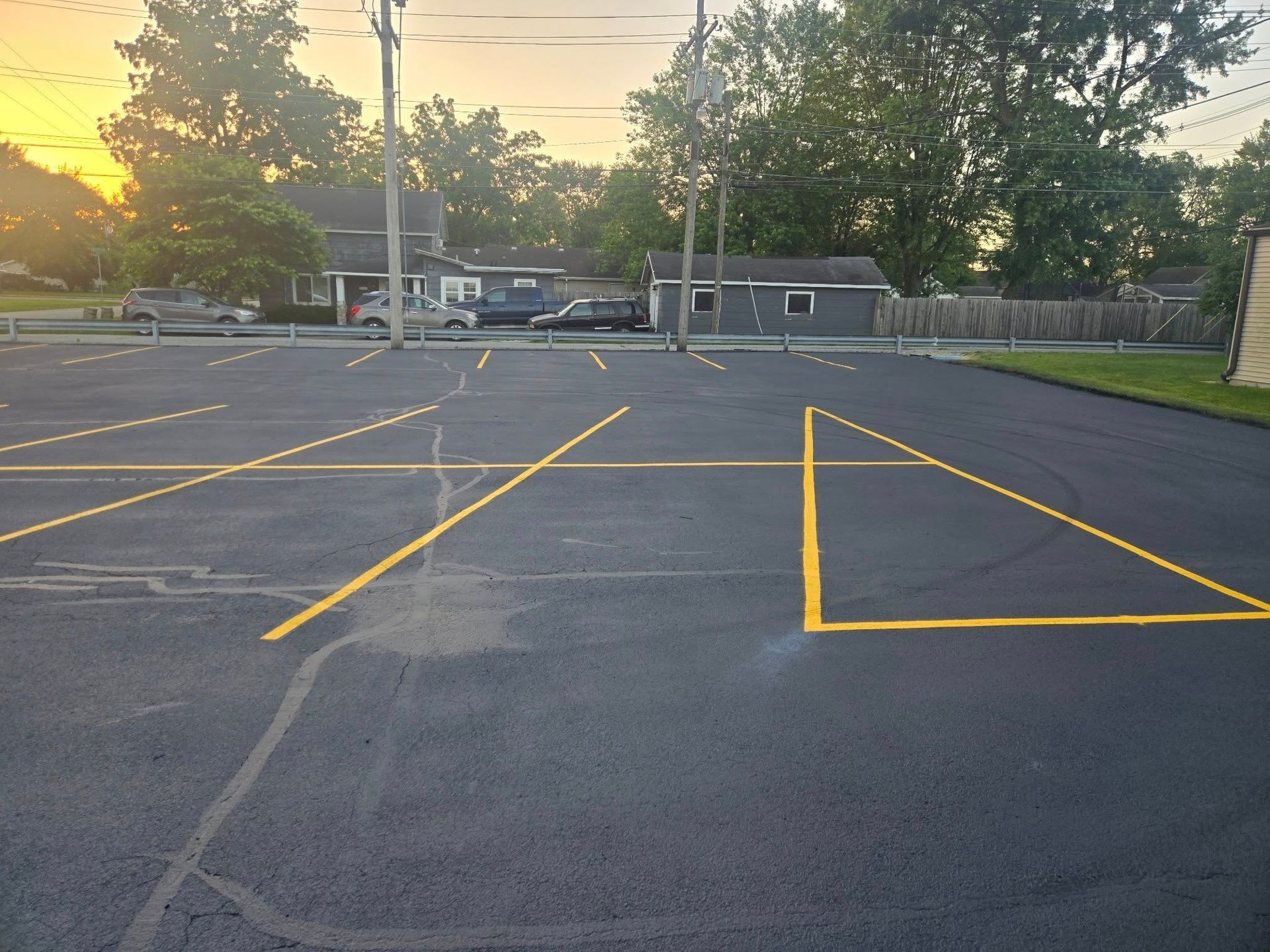 Empty parking lot with yellow painted lines at sunset, trees and buildings in the background.