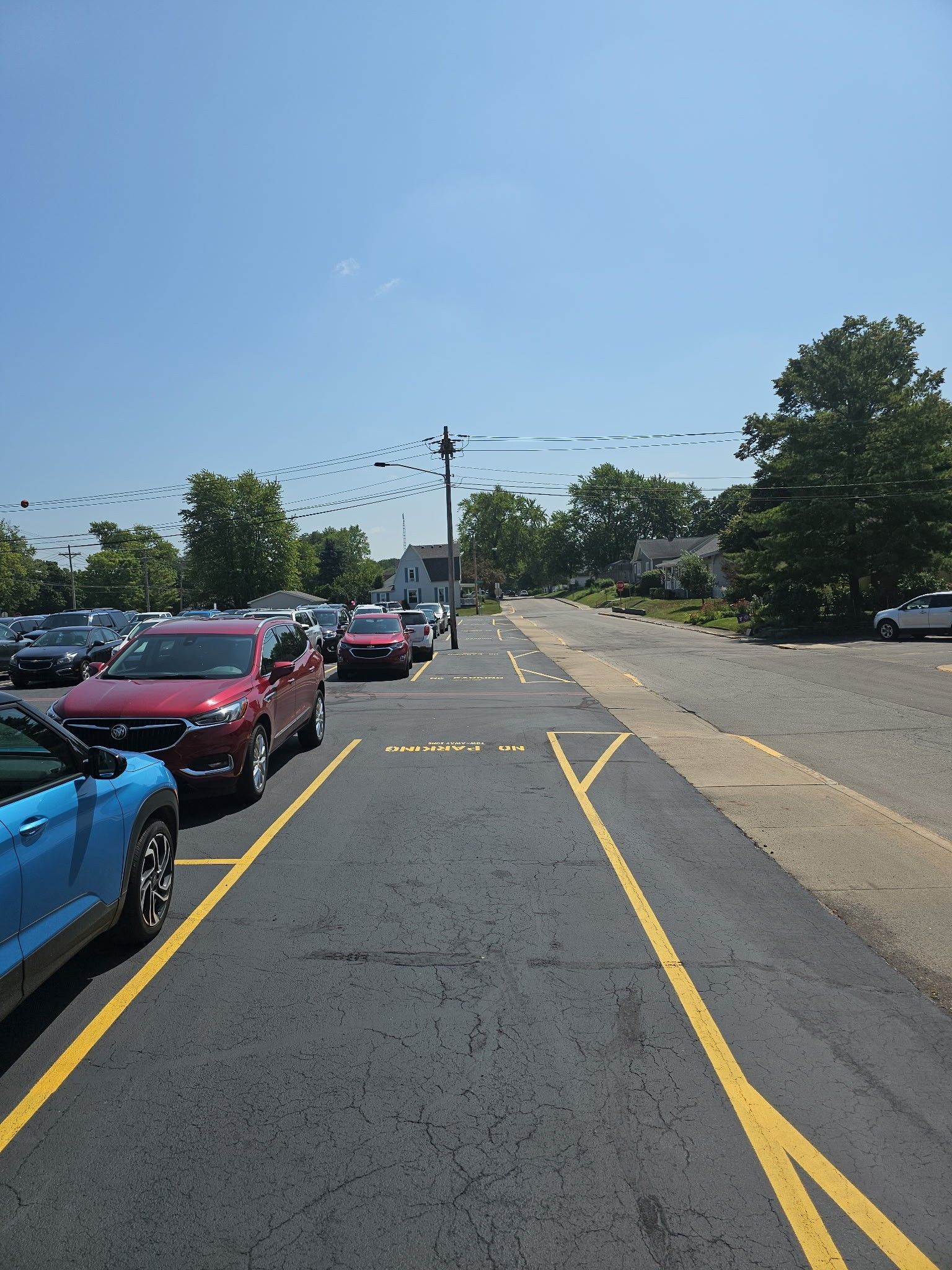 Parking lot with cars lined along the left, a blue car in front, and a road extending ahead under a clear sky