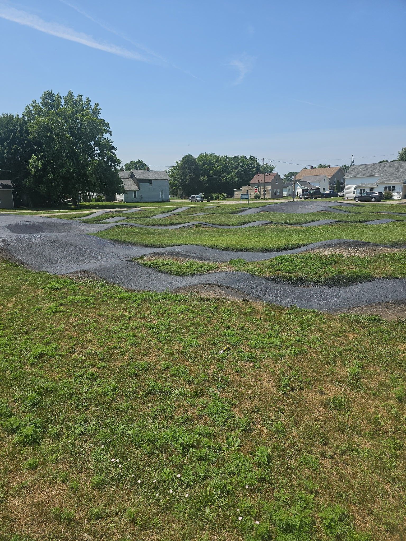 Gravel paths winding through a green park with trees and houses under a clear blue sky