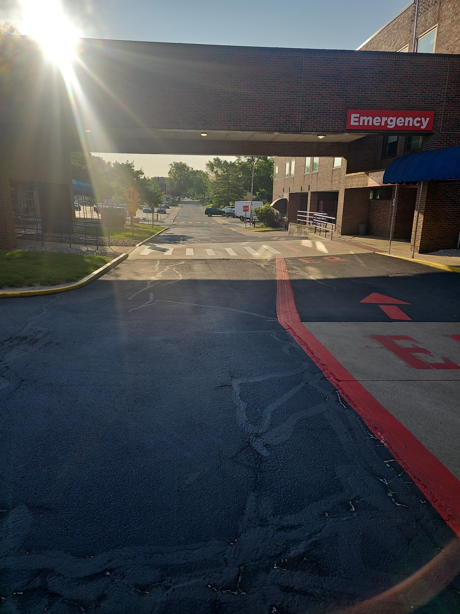 Hospital emergency entrance under bright sunlight, with red directional markings on the pavement.