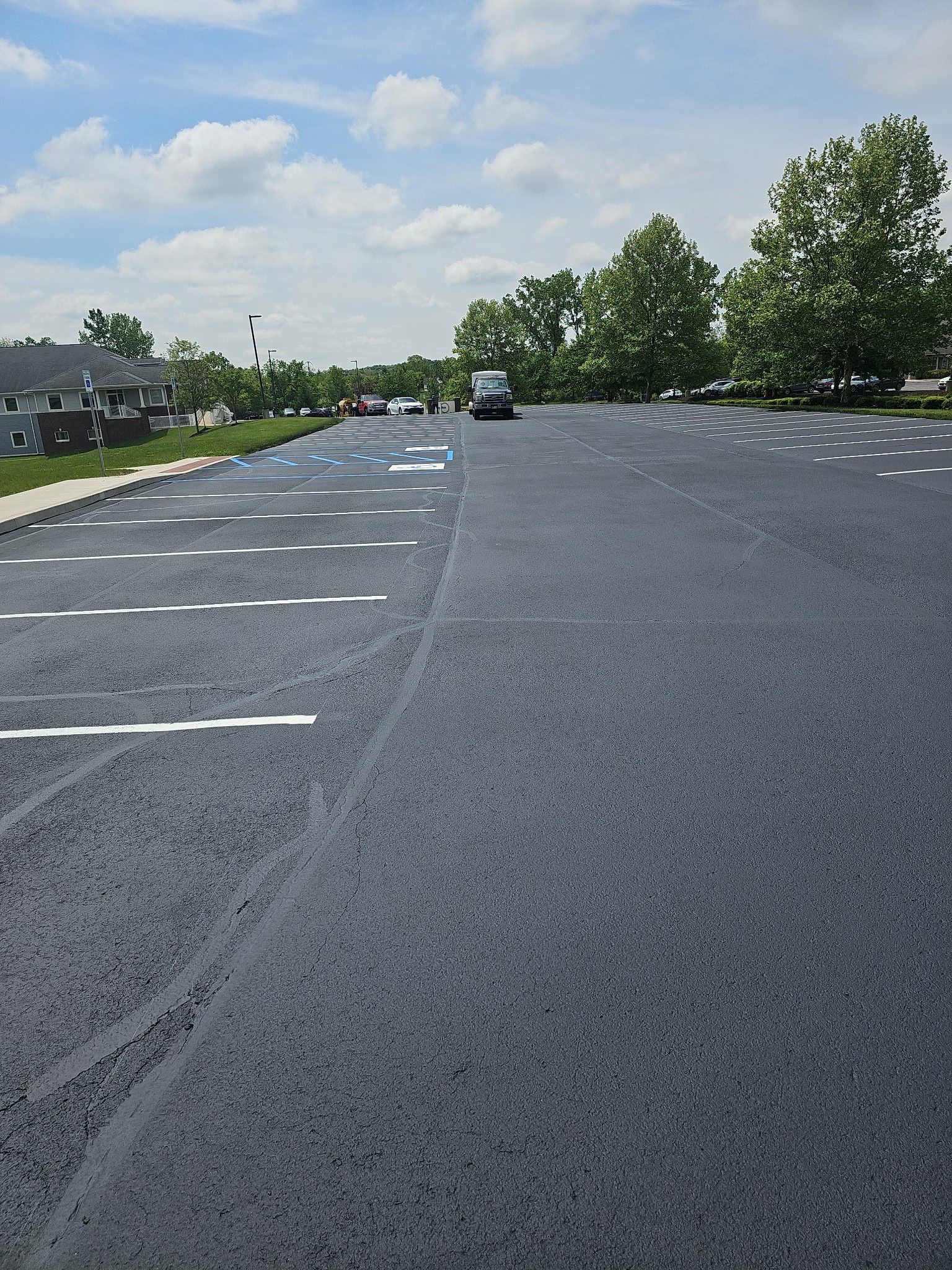 Empty asphalt parking lot with white lines, trees, and parked cars under a partly cloudy sky
