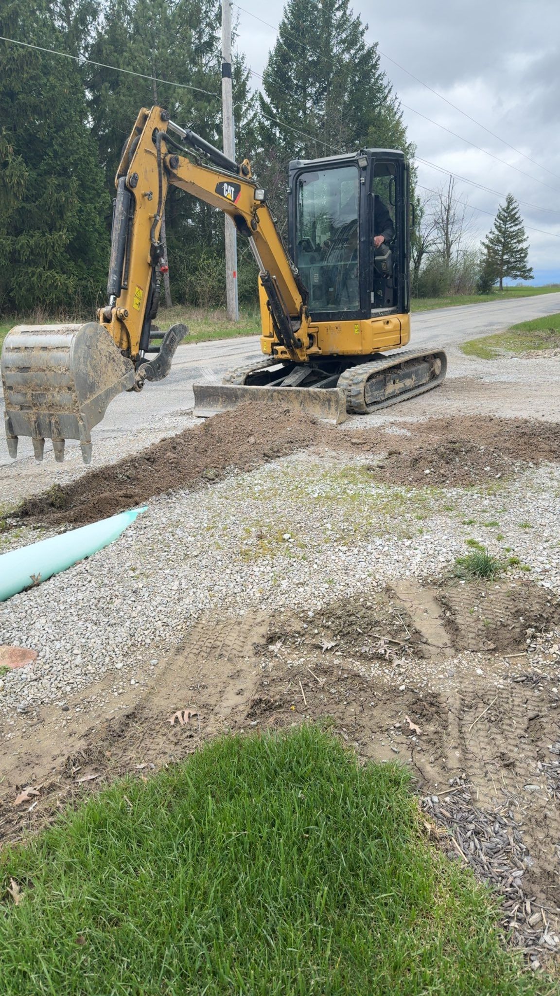 Yellow excavator on gravel near a trench and green grass under cloudy sky