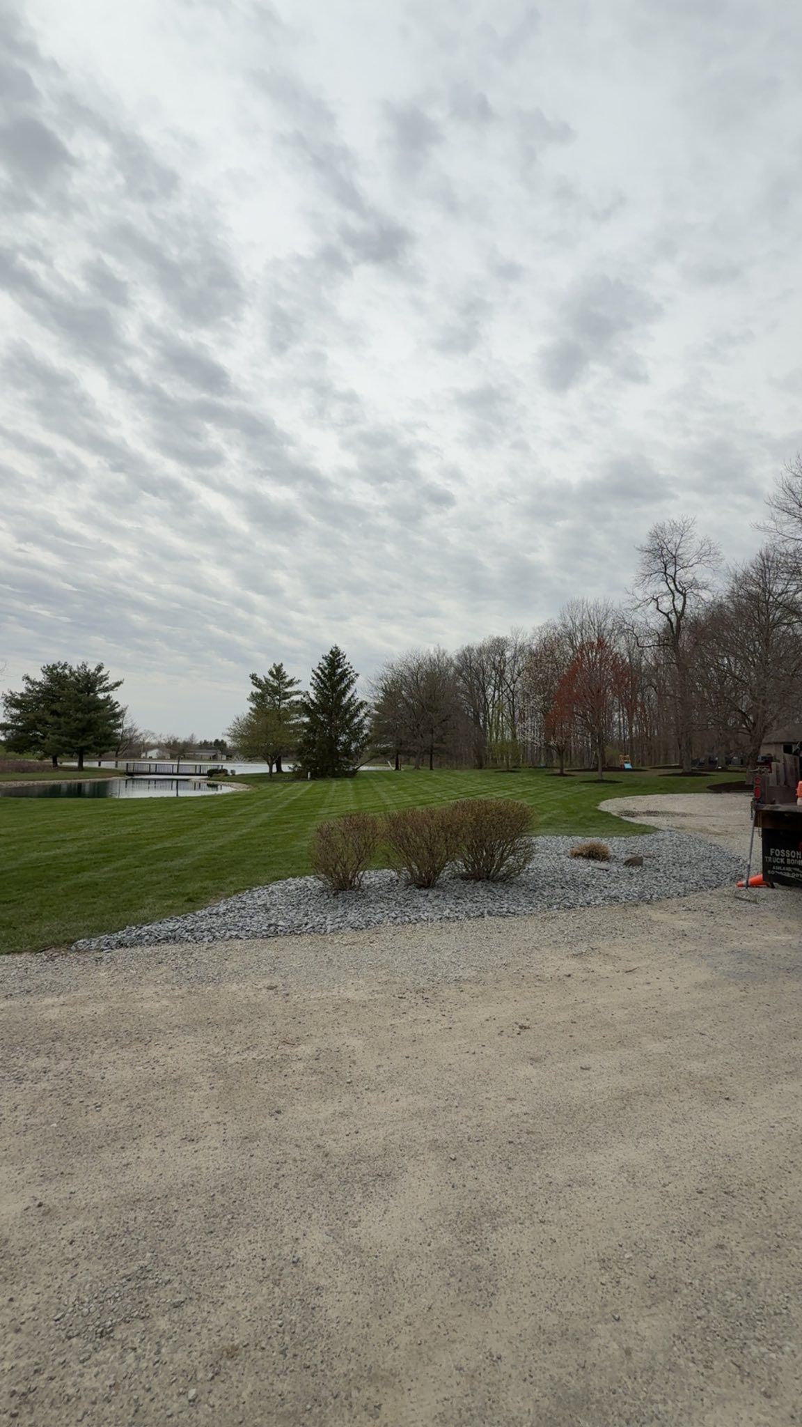 Gravel lot beside a grassy field and trees under a cloudy sky