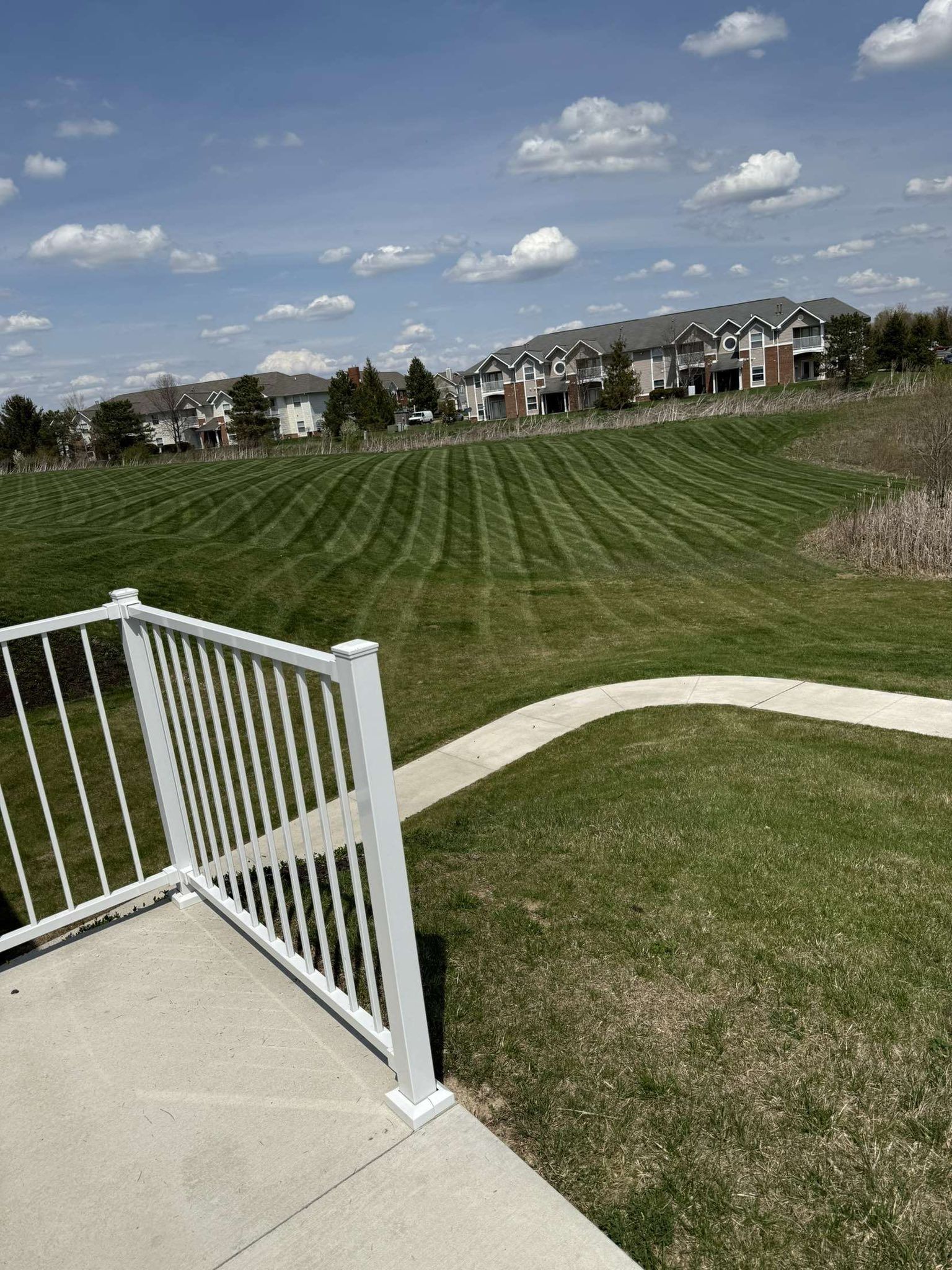 Suburban grassy field with curved sidewalk and white gate, houses in the distance under a cloudy sky