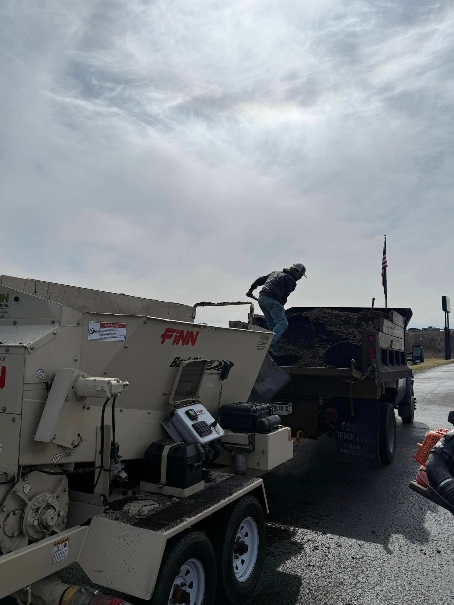 Workers loading a white FMW trailer into a dump truck on a roadside under cloudy skies