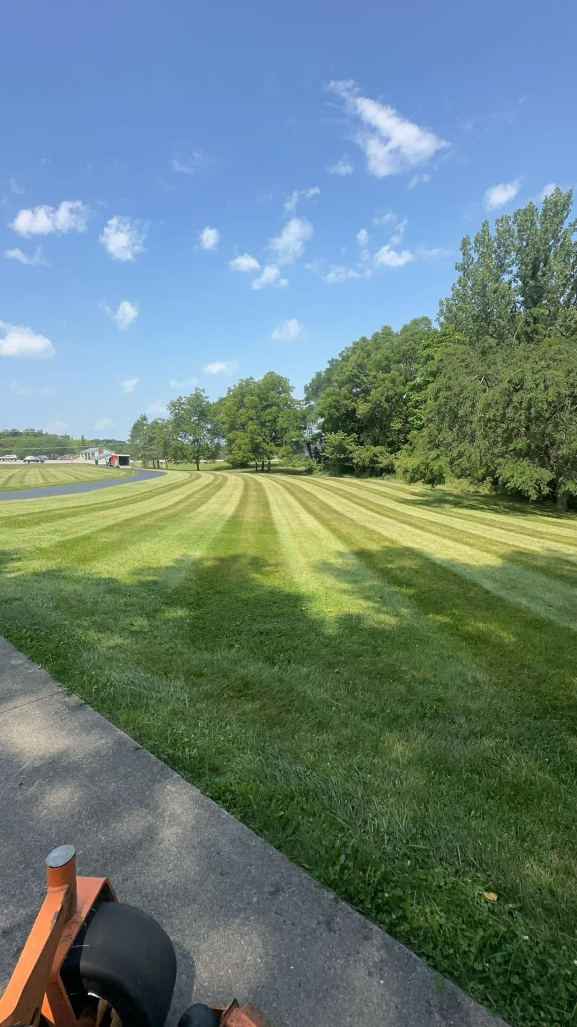 Sunlit mowed field with trees lining the edge beside a paved path under a blue sky