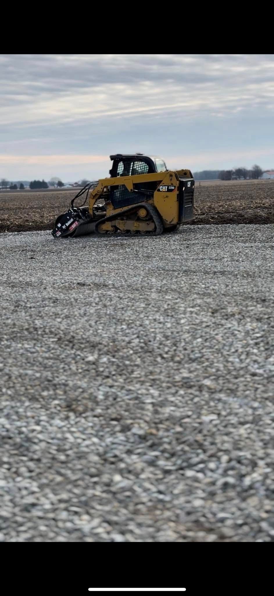 Yellow skid steer clearing a snowy gravel field under a cloudy sky