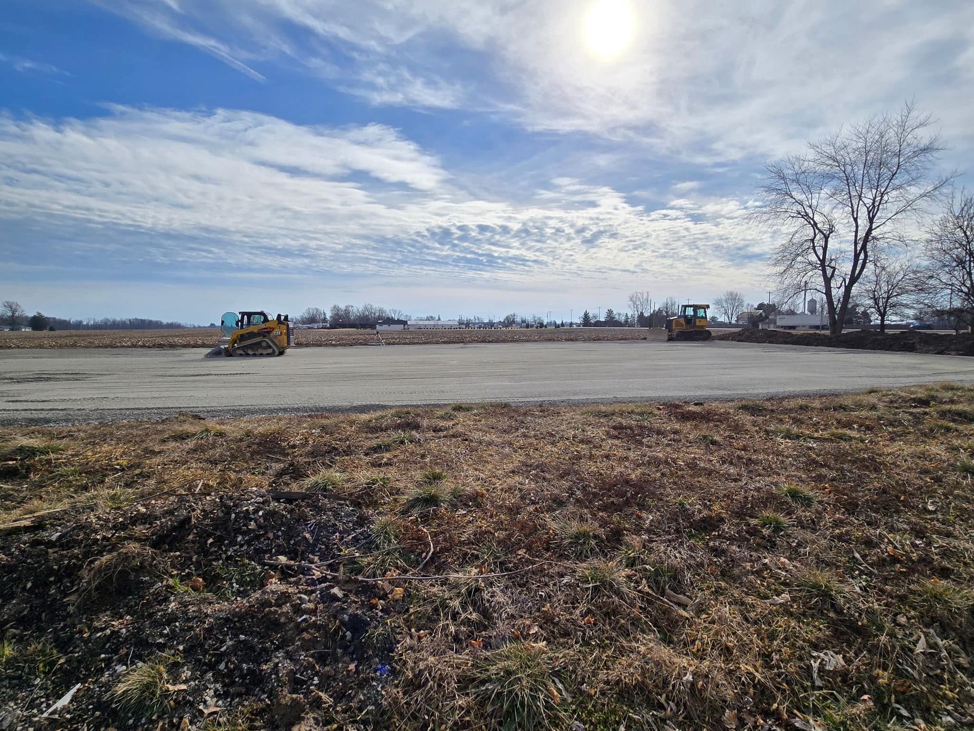 Construction site with a bulldozer under a bright, cloudy sky, framed by grassy ground and bare trees