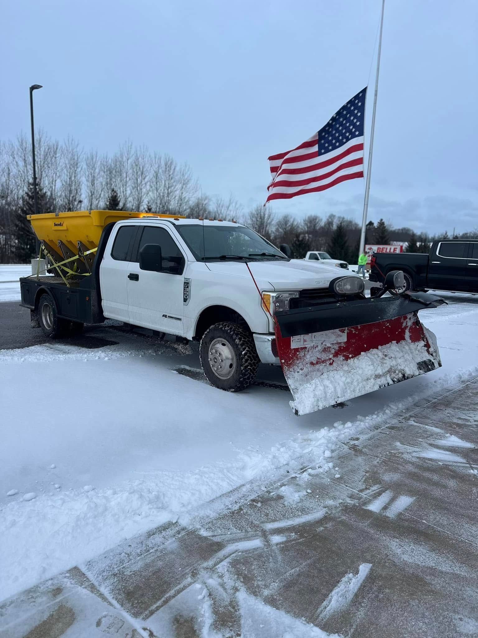 White snowplow truck with red plow and American flag on snowy roadside