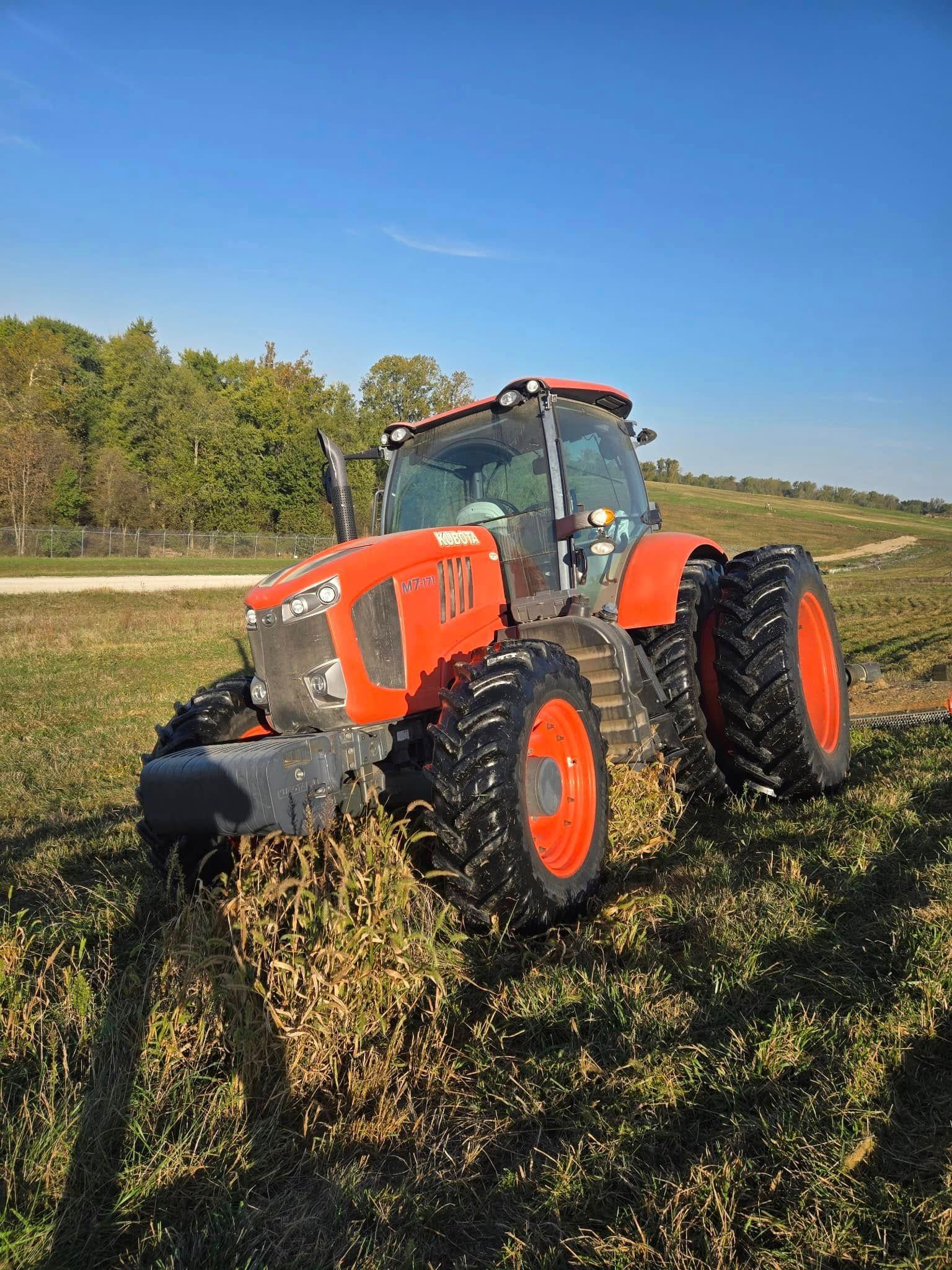Orange tractor plowing a grassy field beside a wooded area under a clear blue sky