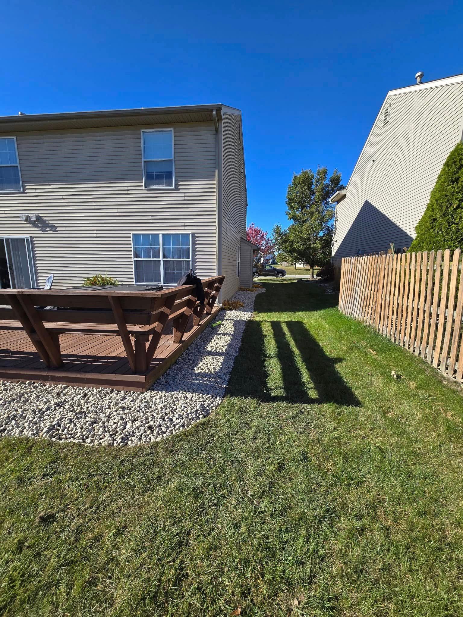 Backyard with a wooden deck, green lawn, gravel path, house siding, and a fence under a clear blue sky