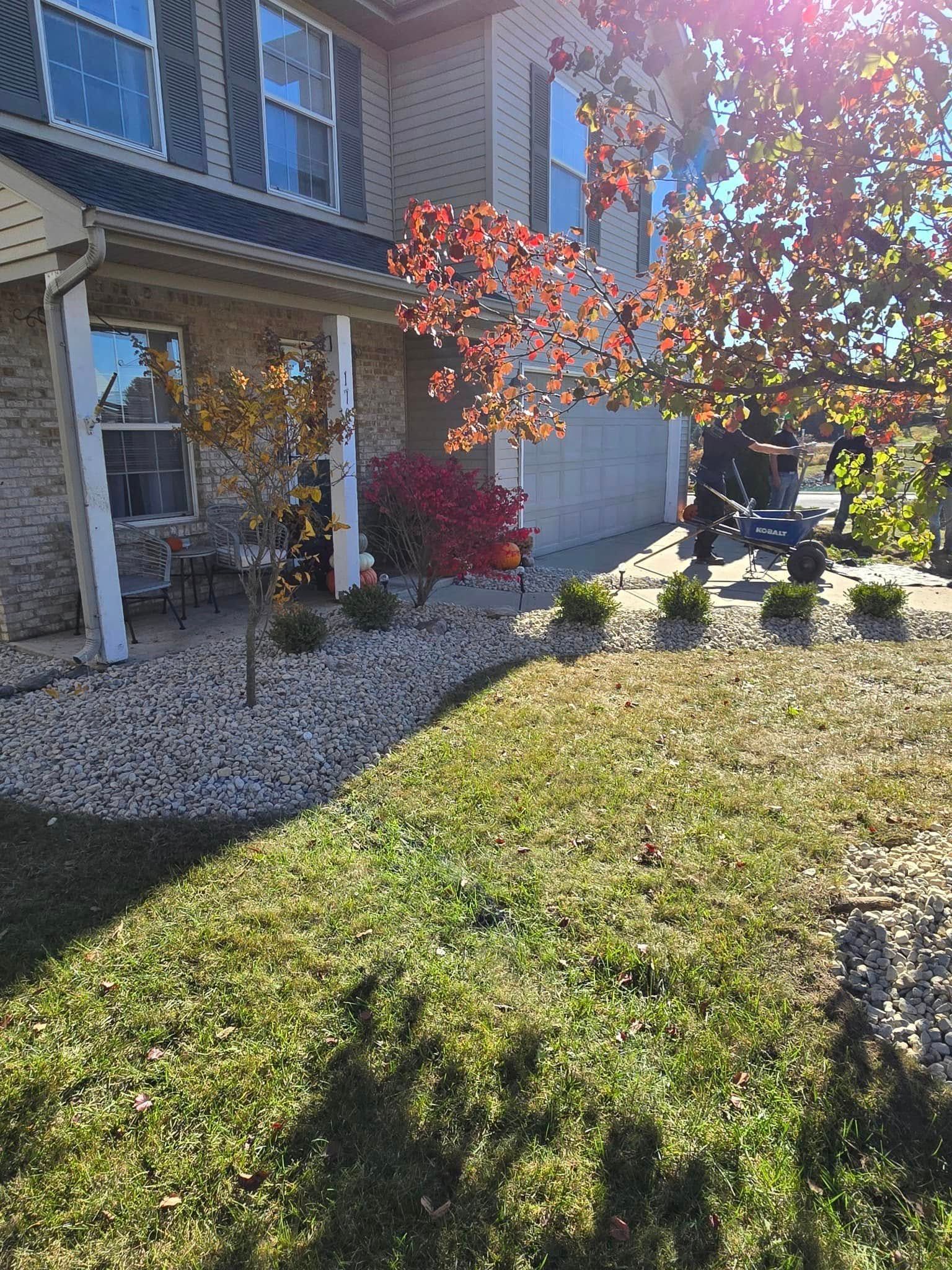 Front yard of a house with gravel, green lawn, and a tree with red leaves in bright sunlight