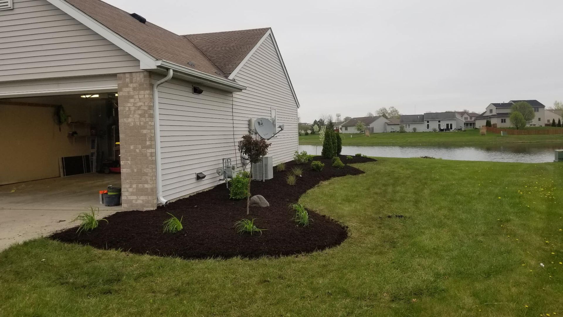 Suburban house with mulched flower bed and young tree beside a lakeside lawn