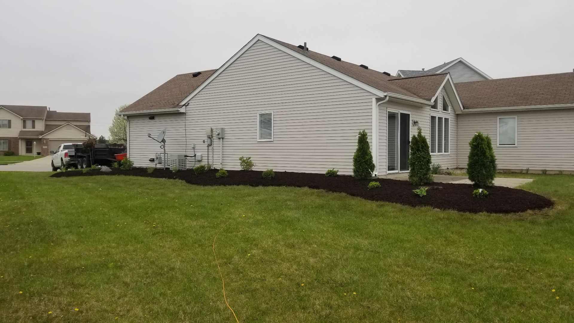Suburban house with light siding, dark mulch landscaping, and a wide green lawn on an overcast day