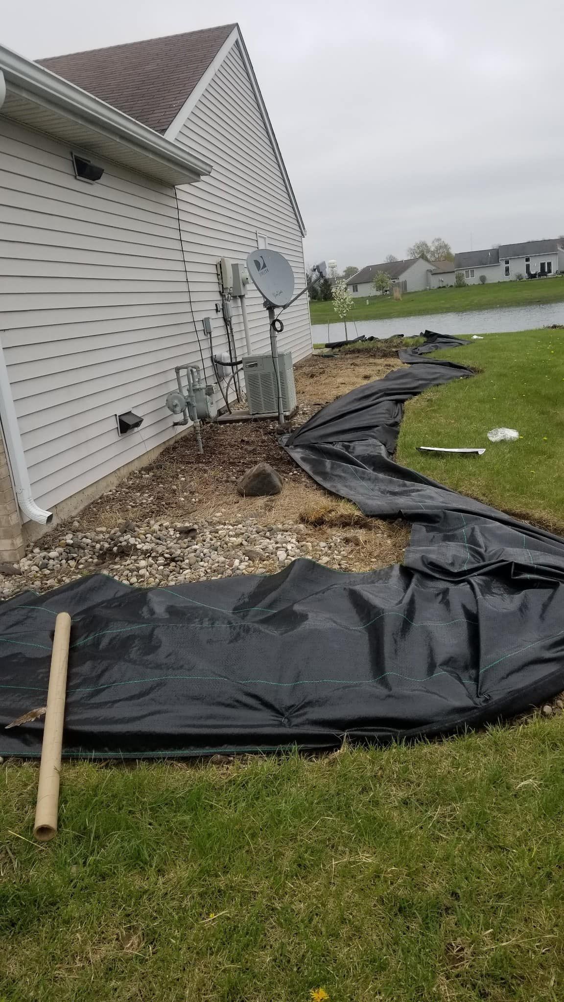 Black landscape fabric laid along a house foundation beside white siding on a grassy yard.