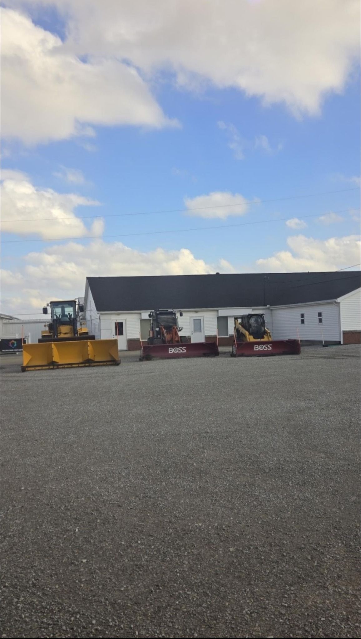 Wide gravel lot with construction equipment in front of a long white building under a cloudy blue sky
