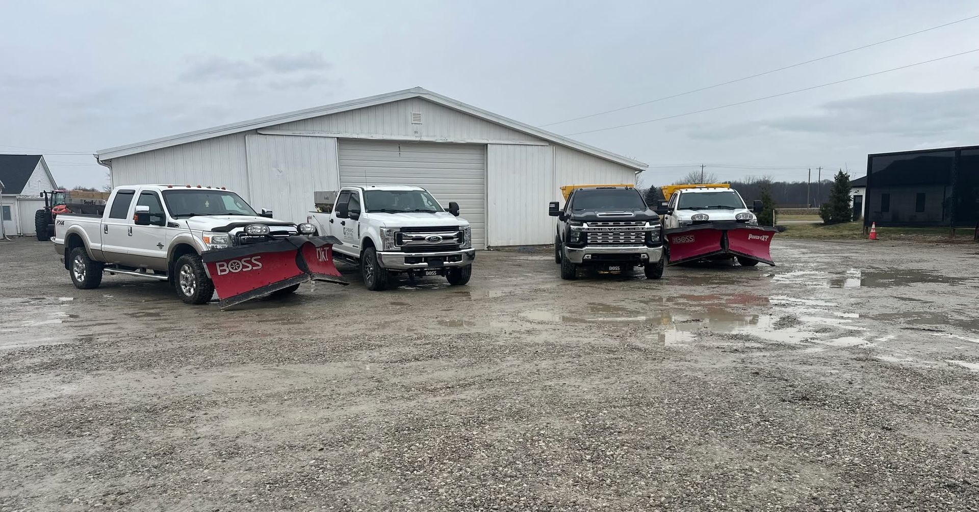 Four pickup trucks parked in a gravel lot in front of a white warehouse on an overcast day