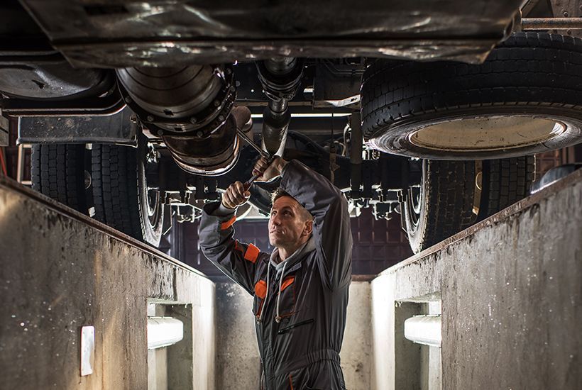 Mechanic inspecting a vehicle’s underside in a narrow repair bay, wearing a hood.