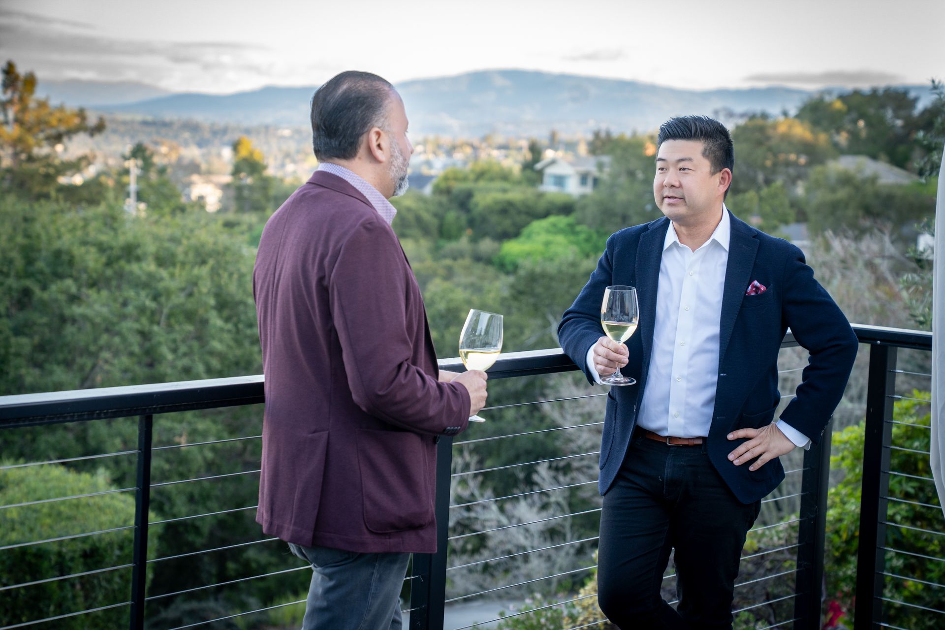 Two men are standing on a balcony holding wine glasses and talking to each other.