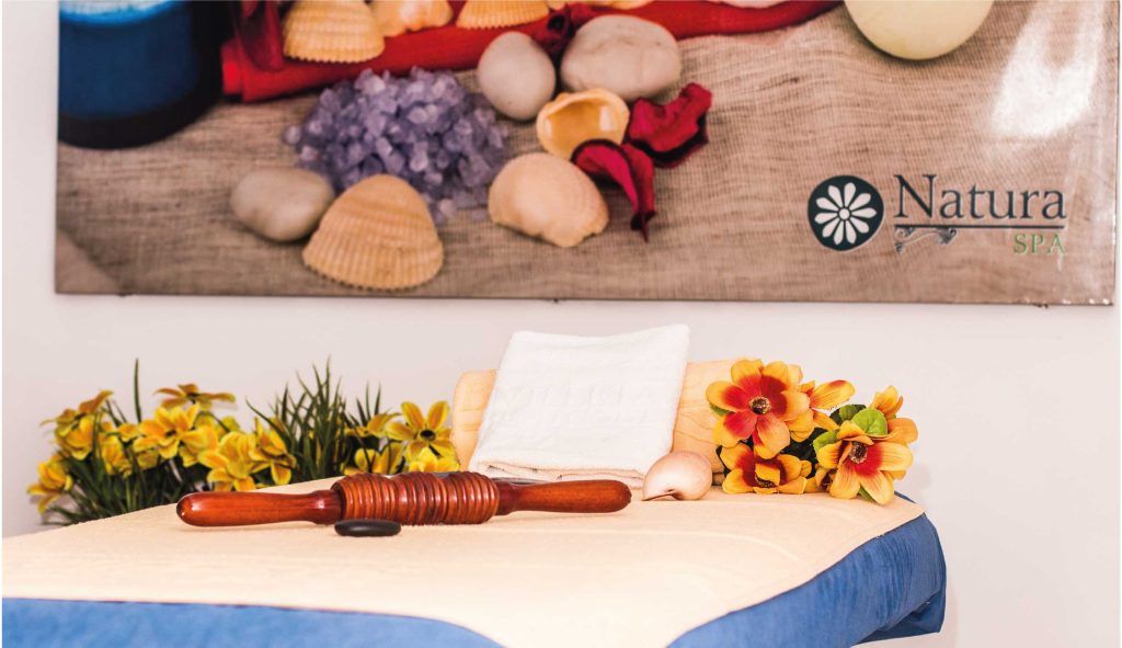 Mesa de spa con flores, velas, piedras y toallas dobladas bajo una pancarta de Natura.