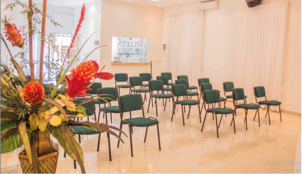 Sala de conferencias con filas de sillas verdes, un arreglo floral al frente y cortinas color crema.