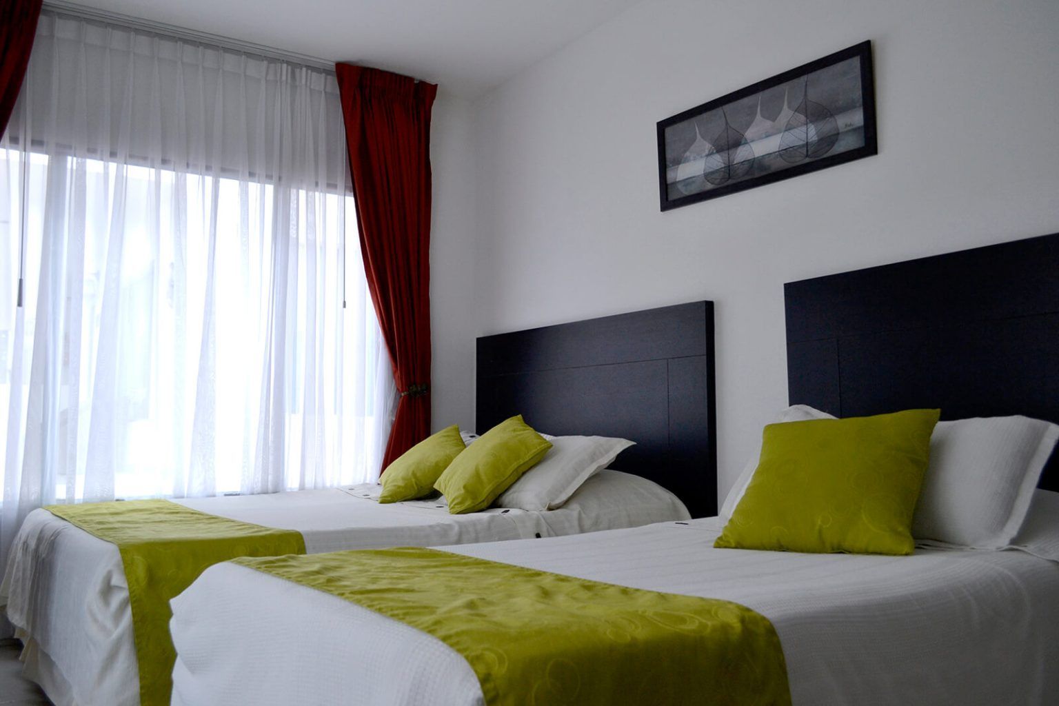 Habitación luminosa con una cama hecha de tela blanca, verde y amarilla, junto a una gran ventana con cortinas rojas.