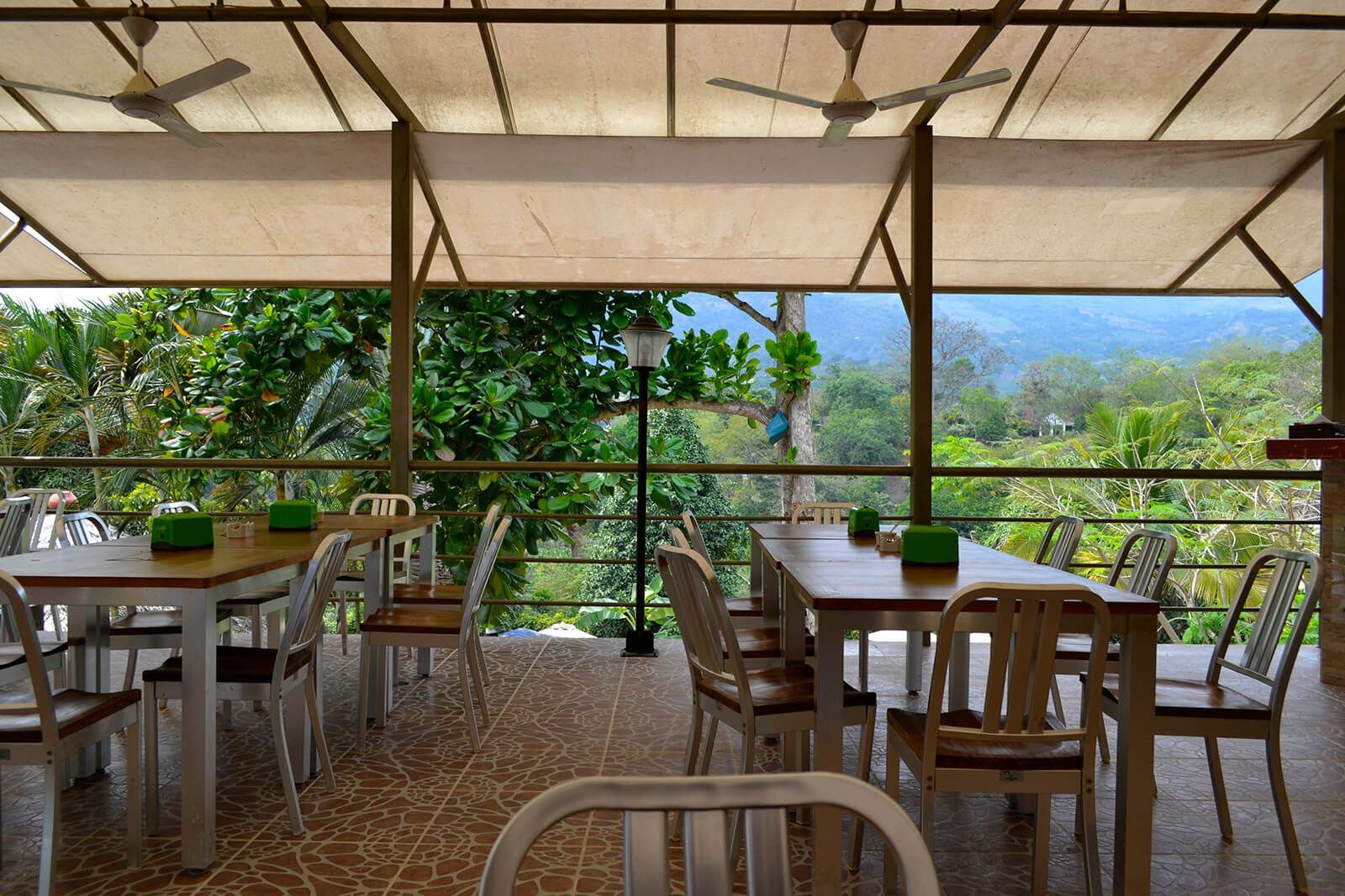 Terraza al aire libre en una cafetería, bajo un toldo, con vistas a frondosos árboles verdes y colinas lejanas.