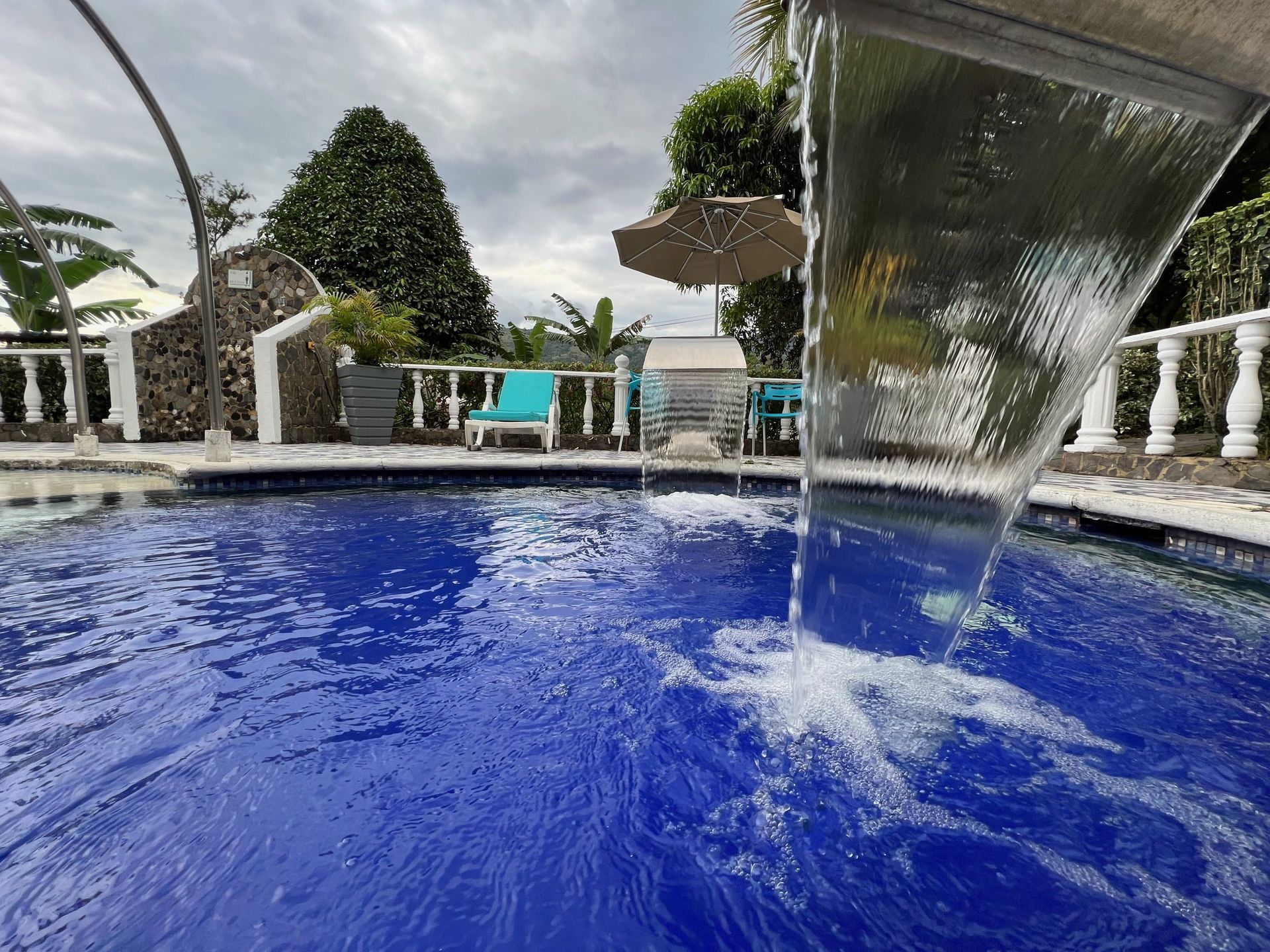 Una fuente junto a la piscina que rocía agua sobre una piscina azul con barandillas blancas y tumbonas al fondo.