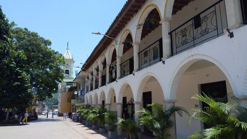 Edificio blanco con arcos y balcones junto a un paseo arbolado bajo un cielo azul.
