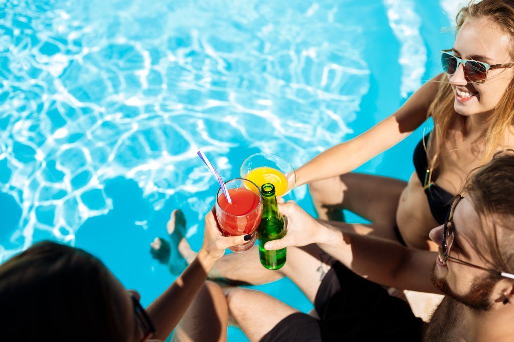 Un grupo de amigos brinda con coloridos cócteles junto a una piscina de un azul brillante en un día soleado.
