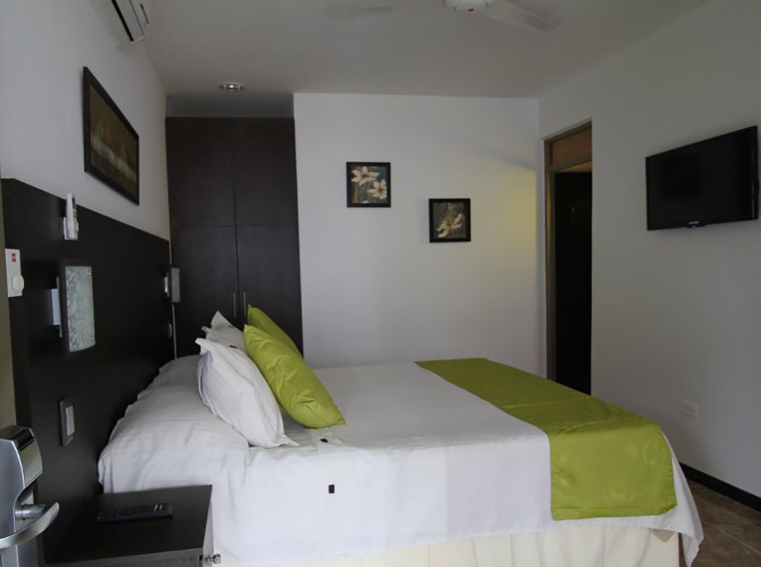 Habitación de hotel moderna con cama blanca, detalles en verde, televisor montado en la pared y cuadros enmarcados.