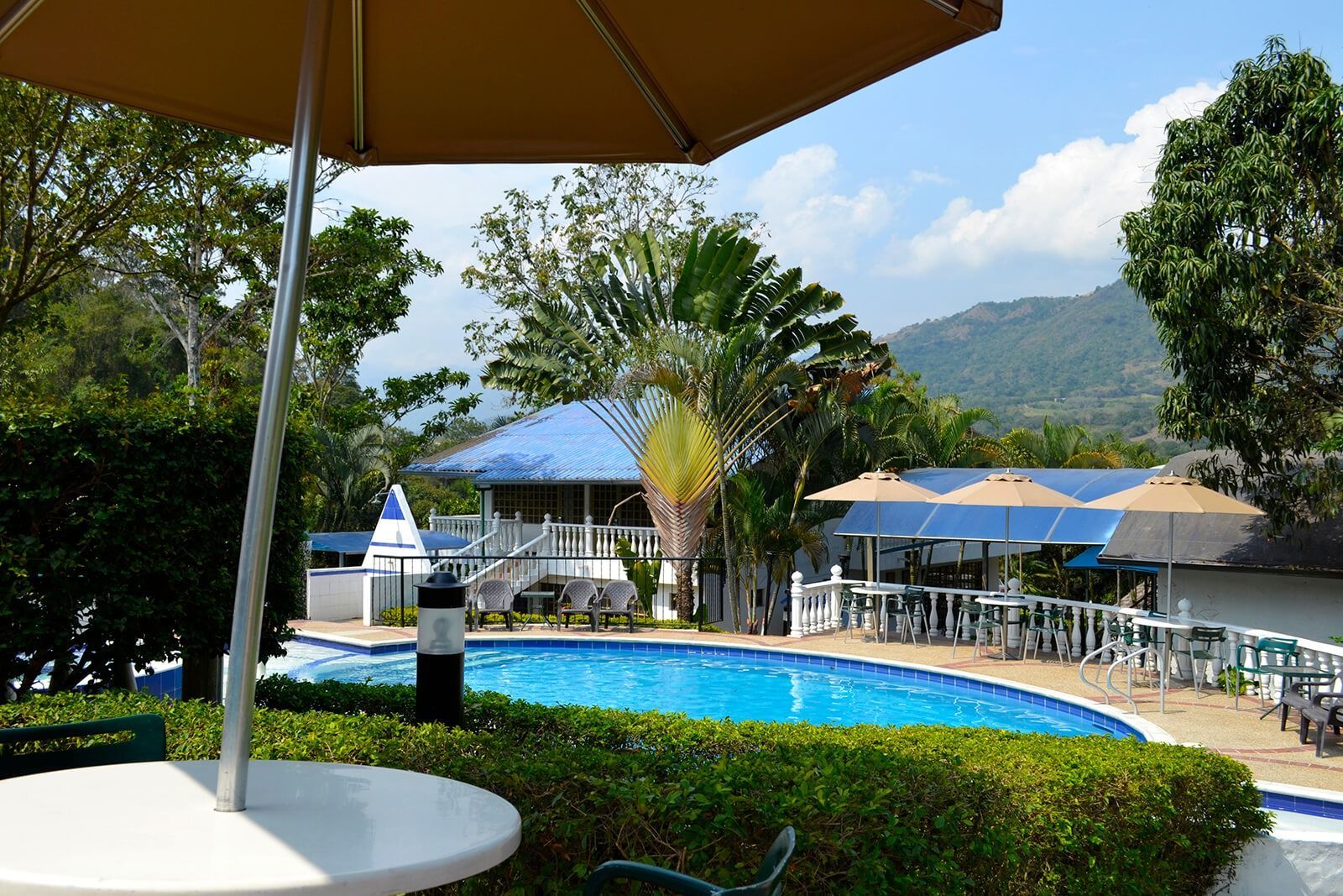 Piscina del complejo turístico con sombrillas azules, sillas blancas y vistas a la montaña bajo un cielo soleado.