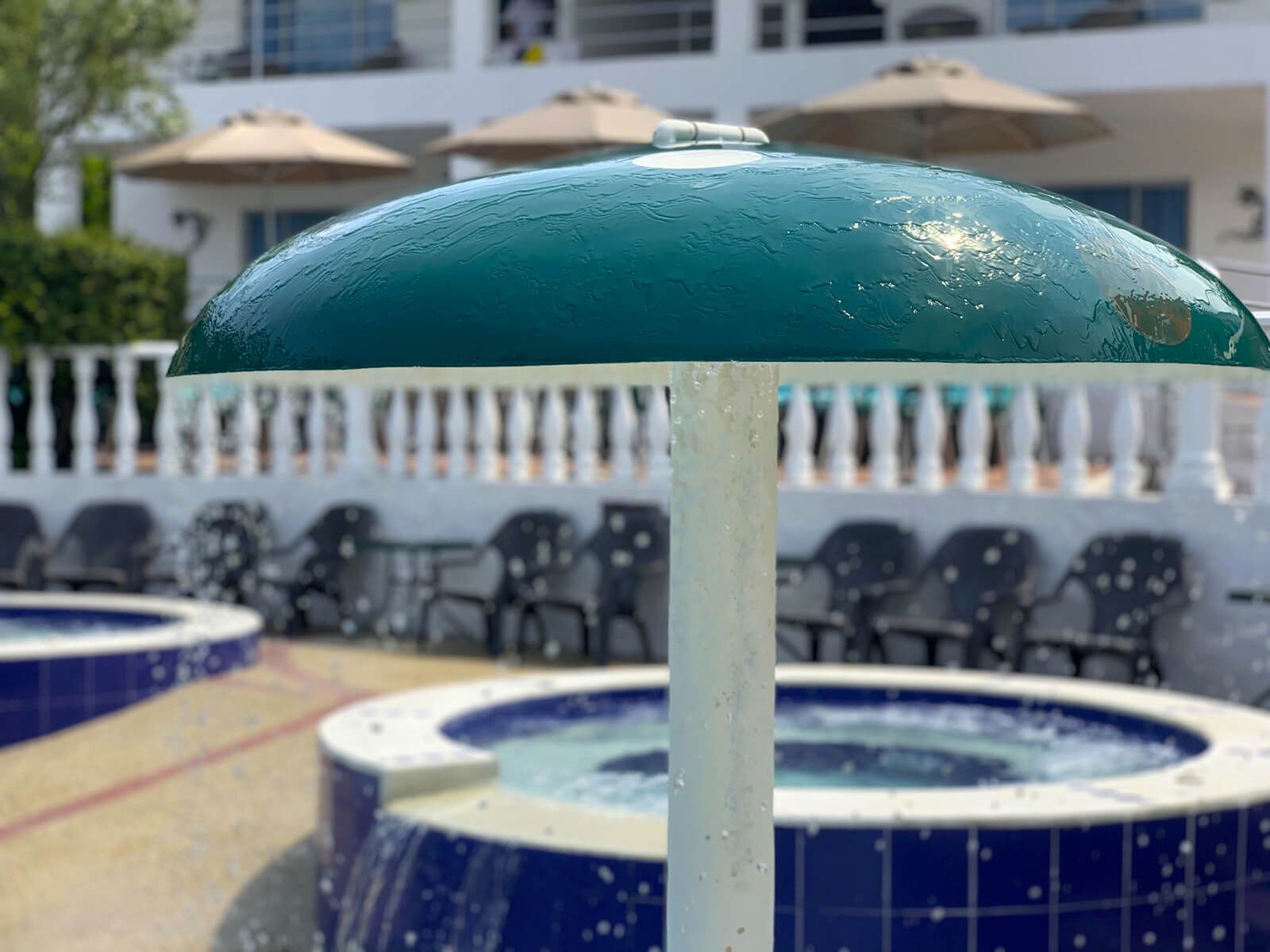 Una sombrilla verde de patio se alza sobre una zona de piscina con azulejos, mesas y sillas azules y blancas.