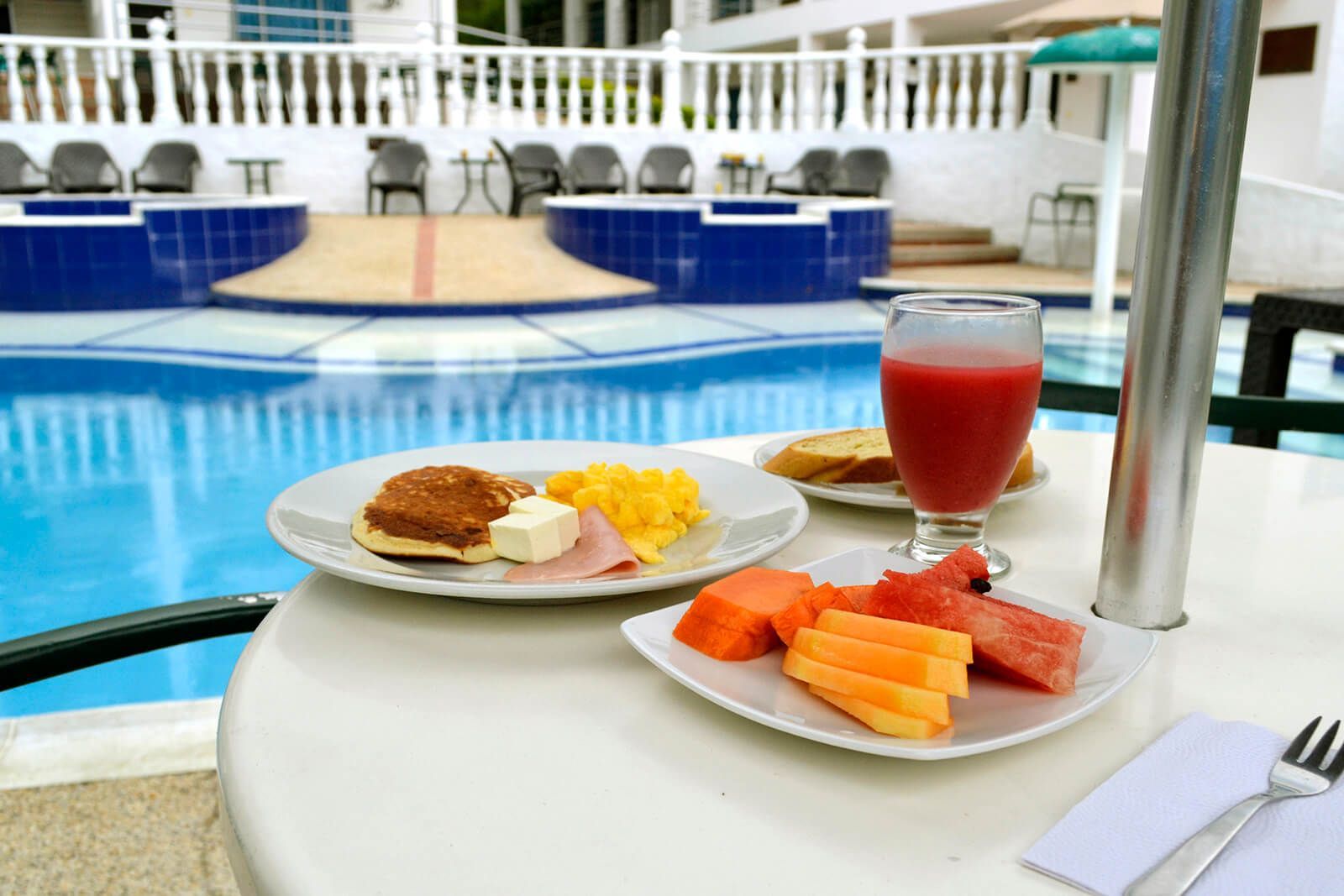 Platos de desayuno y una bebida roja sobre una mesa junto a una piscina azul.