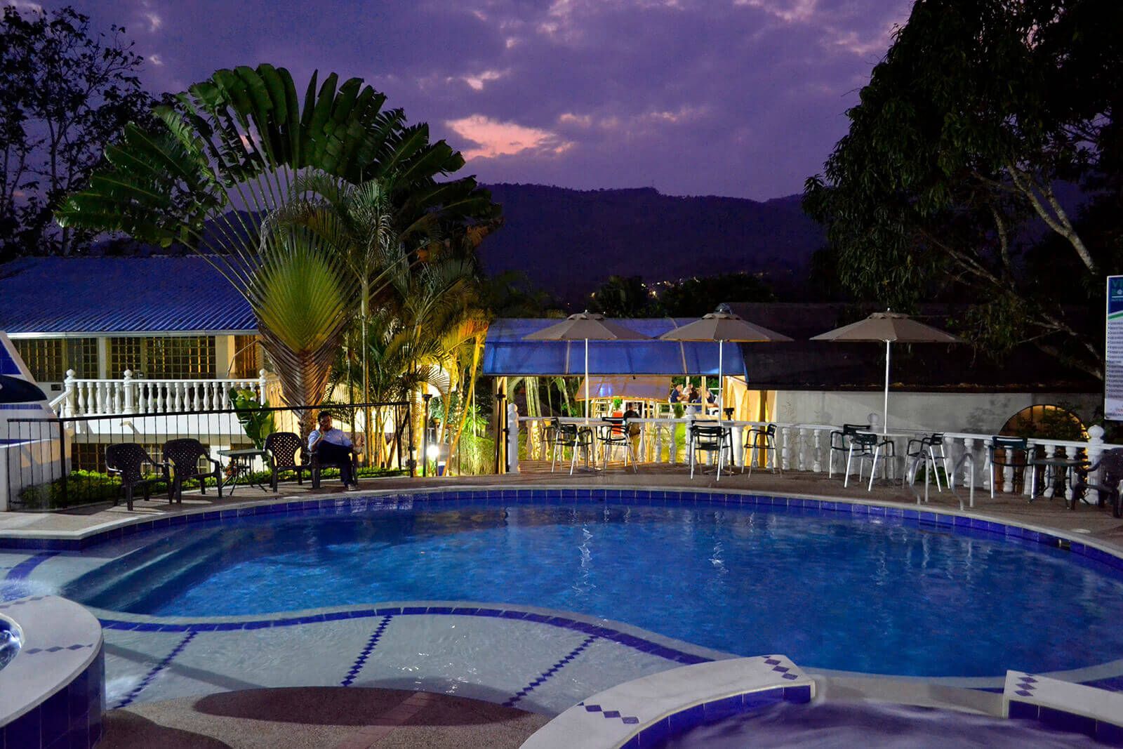 Piscina del resort al atardecer con agua azul, palmeras, cabañas iluminadas y un cielo púrpura.