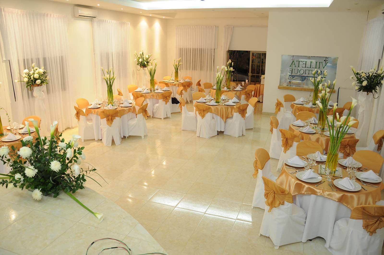 Salón de banquetes decorado con mesas redondas, mantelería blanca y dorada y centros de mesa florales para un evento.