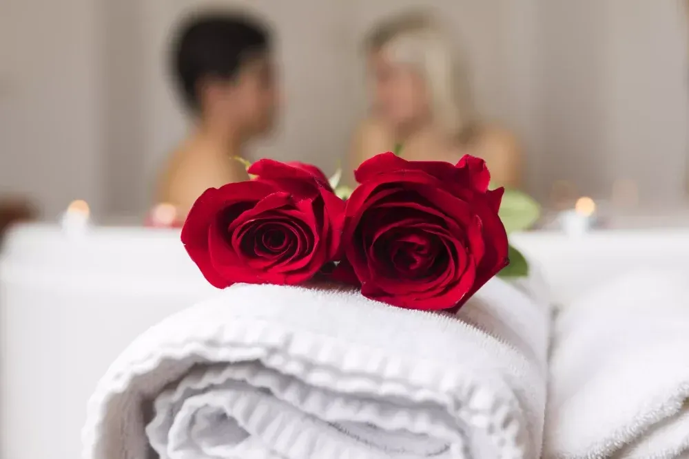 Dos rosas rojas sobre toallas blancas enrolladas en un spa, con una pareja borrosa en una bañera al fondo.