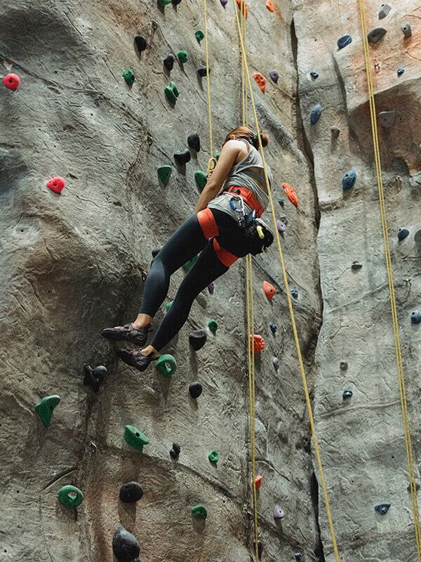 Una persona practica escalada en roca en un interior, en una pared gris con presas de colores y una cuerda de seguridad.